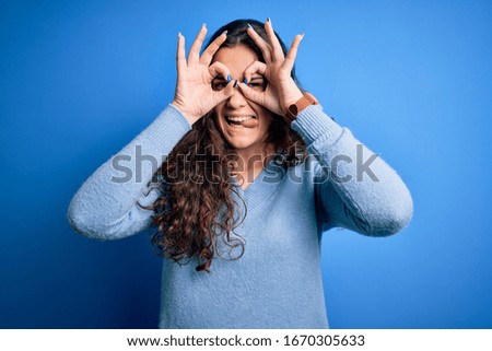 Young beautiful woman with curly hair wearing blue casual sweater over isolated background doing ok gesture like binoculars sticking tongue out, eyes looking through fingers. Crazy expression.