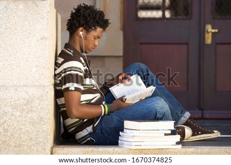 Male student wearing earphones studying books outside building Royalty-Free Stock Photo #1670374825