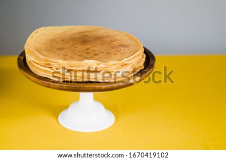 Food presentation photo of Russian pancakes lying on wooden plate with white stand. Yellow, grey colours on background. Image for Eastern Orthodox religion festival Maslenitsa, food blog, wallpaper.