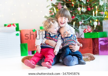 Three happy children - teenager boy, toddler girl and their newborn baby brother - playing together under a beautiful Christmas tree