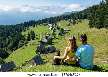 CLOSE UP Young couple observes the rural mountain landscape with their adorable little dog. Unrecognizable tourist couple hiking around Pokljuka with their puppy look at scenic landscape and cabins Royalty-Free Stock Photo #1670465326