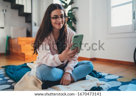 Attractive young woman blogger in casual clothes sitting on floor in modern living room using smartphone writing new post social networks, happy hipster girl chatting online with friends typing text 