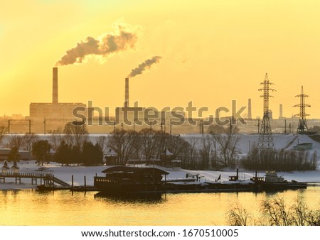 High pipes of a heat station in the city of Novosibirsk, power line supports, a railway embankment. Snow on the beach, barges at the pier, bright yellow sunset sky. Without people, space for text