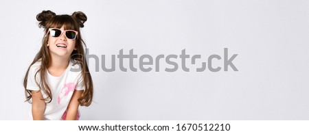 A little smiling girl in sunglasses in a white Studio. In a white t-shirt with a pink Flamingo. Two bunches and loose hair, horns. Royalty-Free Stock Photo #1670512210