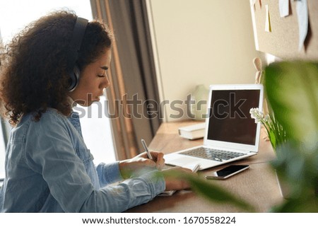 Focused african female student wears headphones elearning studying alone at home office desk. Millennial mixed race teen girl listening audio podcast e learning english language concept making notes. Royalty-Free Stock Photo #1670558224