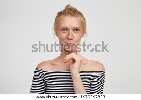 Portrait of young lovely green-eyed redhead female with casual hairstyle twising her mouth while looking thoughtfully at camera and keeping forefinger on her cheek, isolated over white background Royalty-Free Stock Photo #1670567833