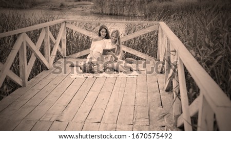 Two cheerful young women have a picnic outdoors on a summer day. Two girlfriends in retro vintage style clothes spend time together on the pond pier.