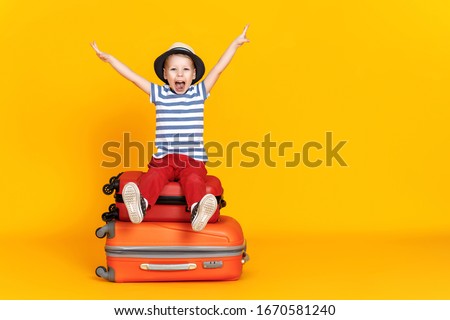 Happy little boy with outstretched arms and open mouth sitting crossed legged on luggage and pretending to fly against yellow background
