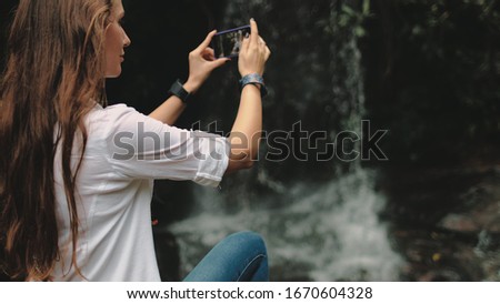 Young Woman Traveler near Waterfall at Rainforest. Caucasian Girl make Video of Mountain Cascade Stream. Female Hiker Holding Smartphone. Exotic Thailand Vacation.