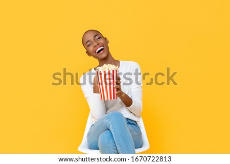 Happy young African American woman laughing while watching movie with popcorn in hand isolated on colorful yellow background