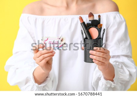 Woman with business card of makeup artist and cosmetics on color background, closeup