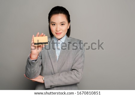 smiling young businesswoman in suit holding credit card on grey background