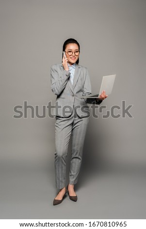 smiling young businesswoman in eyeglasses using laptop and talking on smartphone on grey background