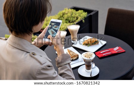Millennial teen girl food travel blogger hold phone take food photo on phone mobile camera sit at cafe table. Young female blogger shoot social media video story blog on smartphone outdoor. Closeup.
