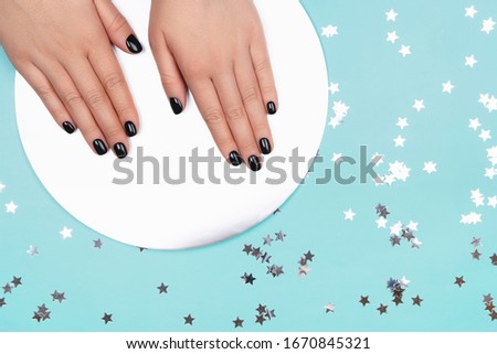 Womans hands with black manicure on silver circle and pastel blue background. Top view.