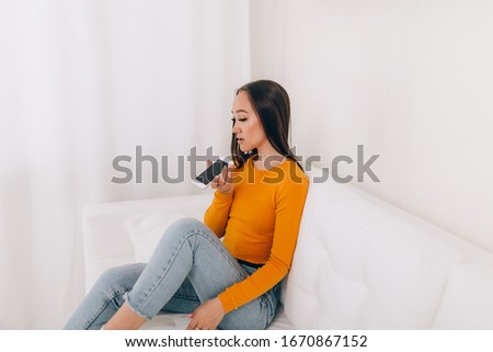 A young girl sitting on sofa records an audio message on the phone in an orange sweater on white sofa in the business room