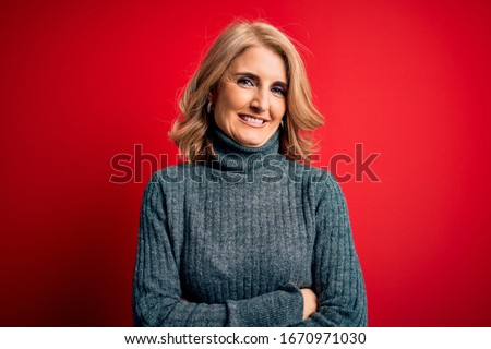 Middle age beautiful blonde woman wearing casual turtleneck sweater over red background happy face smiling with crossed arms looking at the camera. Positive person.