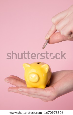 Hands holding yellow money piggy bank and coin in other hand with pink background. Going to throw coin inside piggy bank.