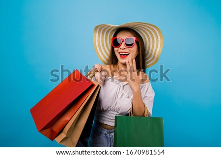 Portrait of happy Asian pretty girl wearing hat and sunglasses holding shopping bags look at away with smiley face isolated over blue background, black friday season sale,colorful shopping concept.