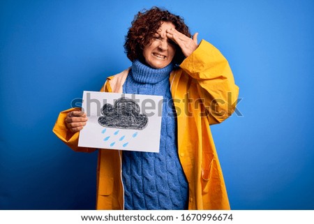 Middle age curly hair woman wearing rain coat holding banner with cloud over blue background stressed with hand on head, shocked with shame and surprise face, angry and frustrated. Fear and upset 