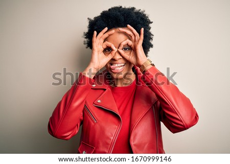 Young beautiful African American afro woman with curly hair wearing casual red jacket doing ok gesture like binoculars sticking tongue out, eyes looking through fingers. Crazy expression.