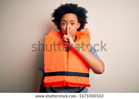 Young African American afro woman with curly hair wearing orange protection lifejacket asking to be quiet with finger on lips. Silence and secret concept.
