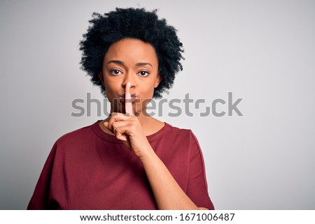 Young beautiful African American afro woman with curly hair wearing casual t-shirt standing asking to be quiet with finger on lips. Silence and secret concept.