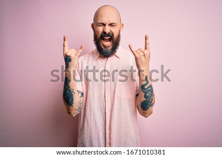 Handsome bald man with beard and tattoo wearing casual shirt over isolated pink background shouting with crazy expression doing rock symbol with hands up. Music star. Heavy concept.