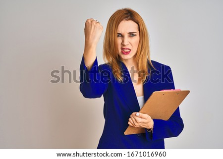 Redhead business caucasian woman holding clipboard over isolated background annoyed and frustrated shouting with anger, crazy and yelling with raised hand, anger concept