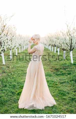 Full-lengh portrait of young blond woman in long elegant dress in the spring apple garden, standing on green grass