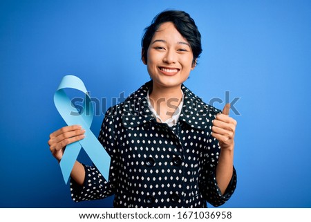 Young beautiful asian girl holding blue cancer ribbon symbol over isolated background happy with big smile doing ok sign, thumb up with fingers, excellent sign