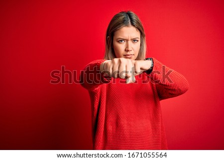 Young beautiful blonde woman wearing casual sweater over red isolated background Punching fist to fight, aggressive and angry attack, threat and violence