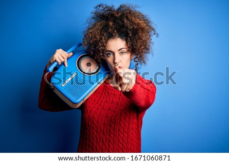 Young beautiful woman with curly hair and piercing listening to music using vintage radio pointing with finger to the camera and to you, hand sign, positive and confident gesture from the front