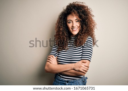 Young beautiful woman with curly hair and piercing wearing casual striped t-shirt happy face smiling with crossed arms looking at the camera. Positive person.