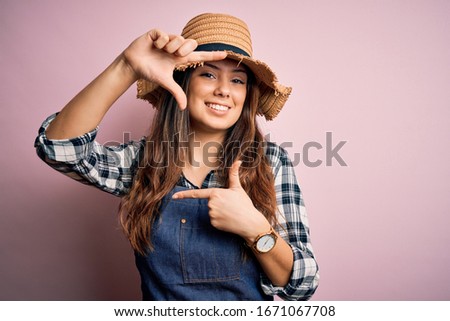 Young beautiful brunette farmer woman wearing apron and hat over pink background smiling making frame with hands and fingers with happy face. Creativity and photography concept.