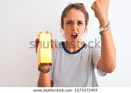 Young beautiful redhead woman holding gold ingot over isolated background annoyed and frustrated shouting with anger, crazy and yelling with raised hand, anger concept