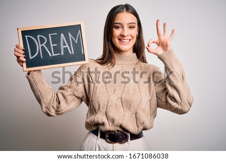 Young beautiful brunette woman holding blackboard with dream message word doing ok sign with fingers, excellent symbol