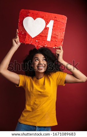 Image of beautiful brunette african american woman with curly hair laughing and holding like symbol placard isolated over red background