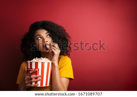 Image of beautiful brunette african american woman with curly hair looking upward and eating popcorn isolated over red background