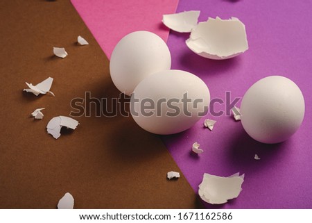 Three white eggs with eggshell pieces on purple, pink and brown colors plain minimal background, angle view, happy Easter day