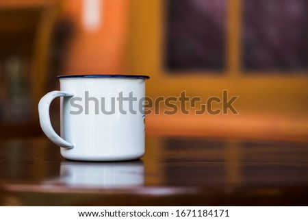 Vintage enamel mug on a classic teak table against simple window background in blur. Antique rustic coffee cup on a brown, smooth wooden surface.