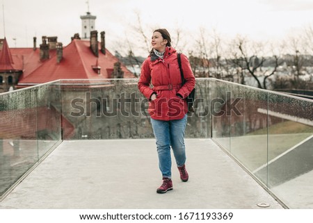 Young brunette girl in a pink jacket and bag walks in the city on the viewing platform with glass railing.