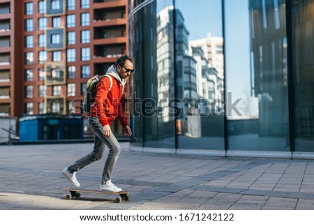 Stylish hipster in red jacket, sunglasses, sneakers and backpack riding on longboard with one foot placed on board and pushing off with the other. Selective focus. Concept of leisure activity, urban.