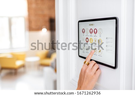 Controlling home with a digital touch screen panel installed on the wall in the living room. Concept of a smart home and mobile application for managing smart devices at home Royalty-Free Stock Photo #1671262936