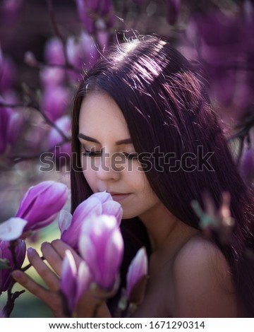 Very close portrait of amazing cute young woman with dark brown long hair inhaling scent of white and pink large magnolia flowers with closed eyes against background of flowering tree on sunny day
