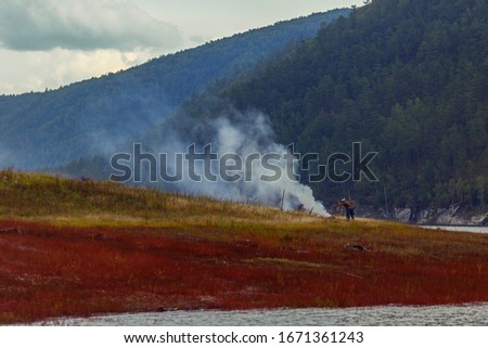A man burns a fire on the shore of a picturesque lake. Smoke from a bonfire in the grass.
