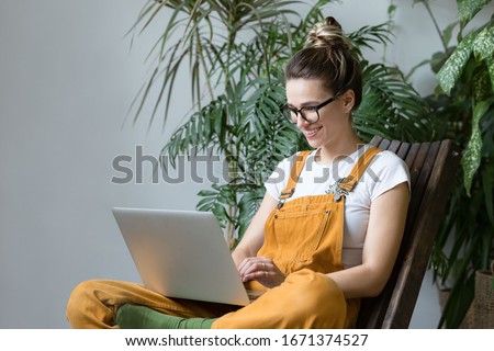 Young female gardener in glasses wearing overalls, sitting on wooden chair in greenhouse, using laptop after work, communicates on Internet with customer. Plant on background.Home gardening, freelance Royalty-Free Stock Photo #1671374527