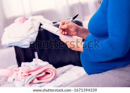 Pregnant woman doing the checklist while packing the suitcase with newborn baby clothes for the hospital.