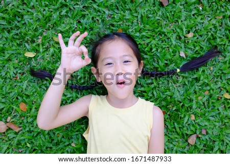 Beautiful asian little child girl making ok sign lying on green grass background. Portrait of smiling kid show confident hands symbol.