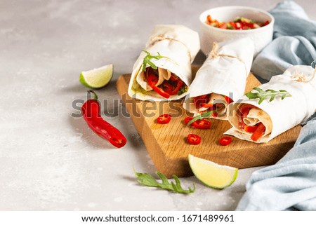 Tortilla wraps sandwiches with vegetables on wooden cutting board with salad, lime and chili pepper. Light grey stone background. 
